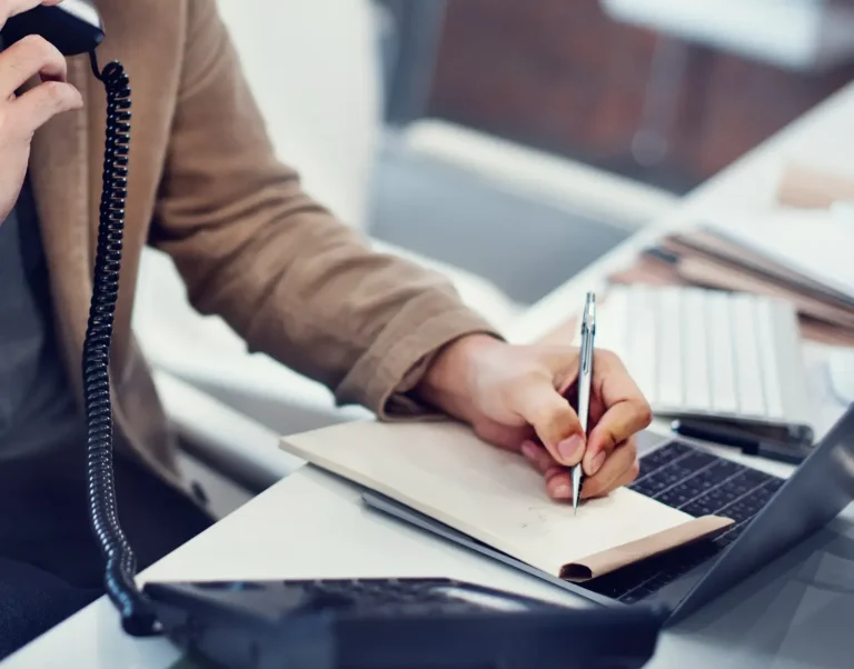 Person am Tisch, telefoniert und macht Notizen in ein Notizbuch. Laptop vorhanden.