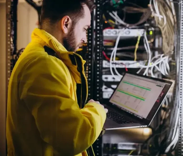 Techniker in gelber Jacke arbeitet an einem Laptop vor Servern.