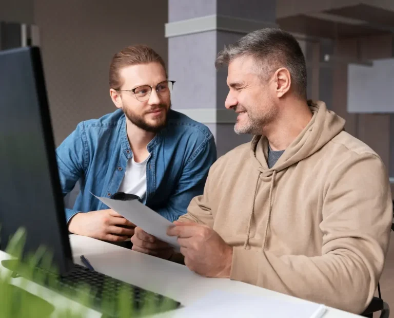 Zwei Männer sitzen am Schreibtisch, schauen auf einen Bildschirm und diskutieren.